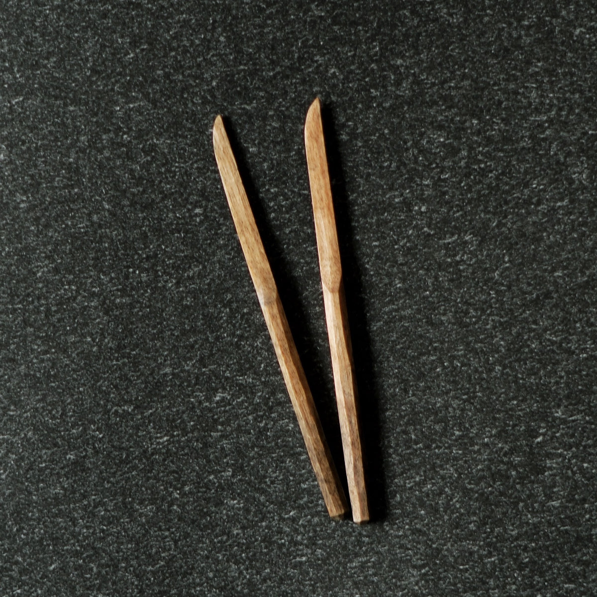 Two slender carved wooden sweet knives with pointed tips, lying on dark speckled stone.