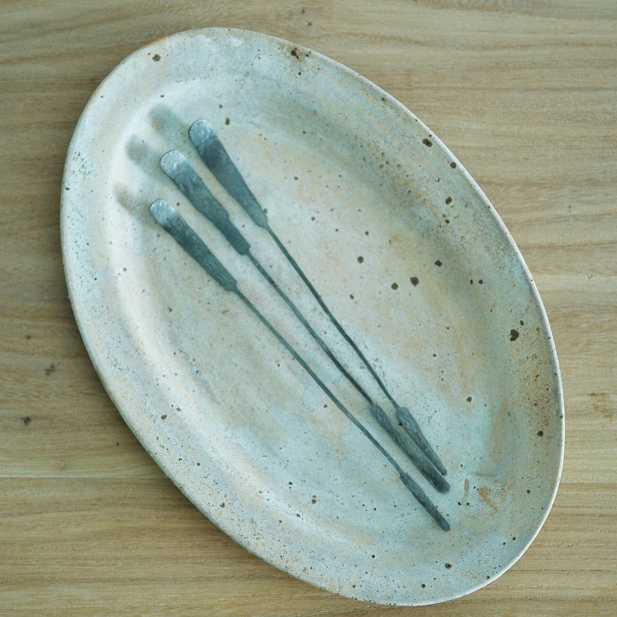 Three long slim tea spoons with pale blue tips, laid diagonally across a speckled oval plate.