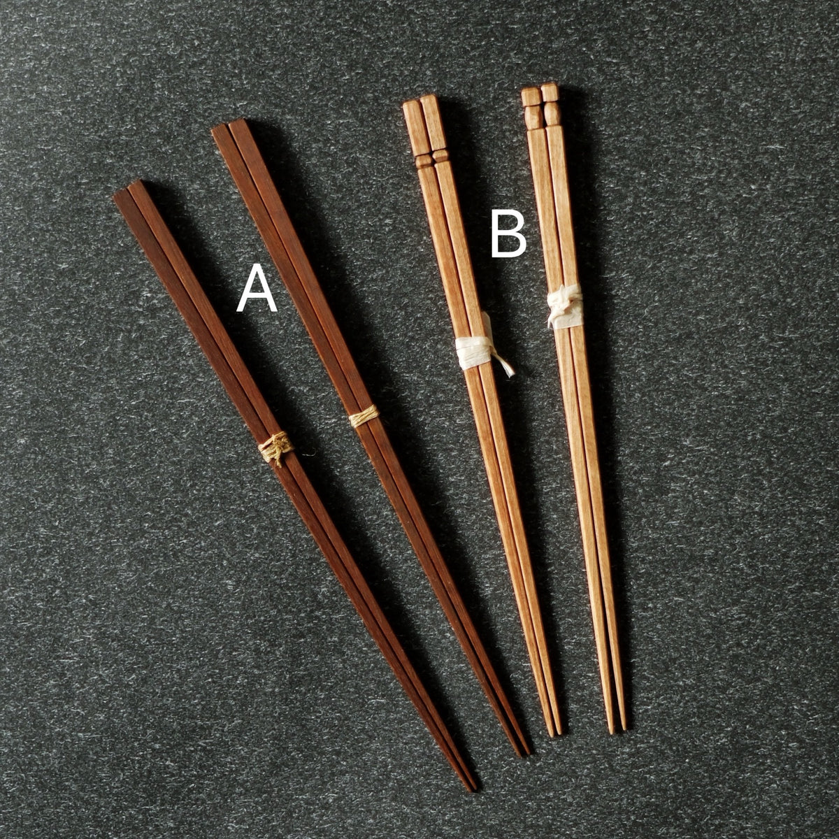 Two pairs of wooden chopsticks marked A and B, one dark and one pale, on dark speckled stone.