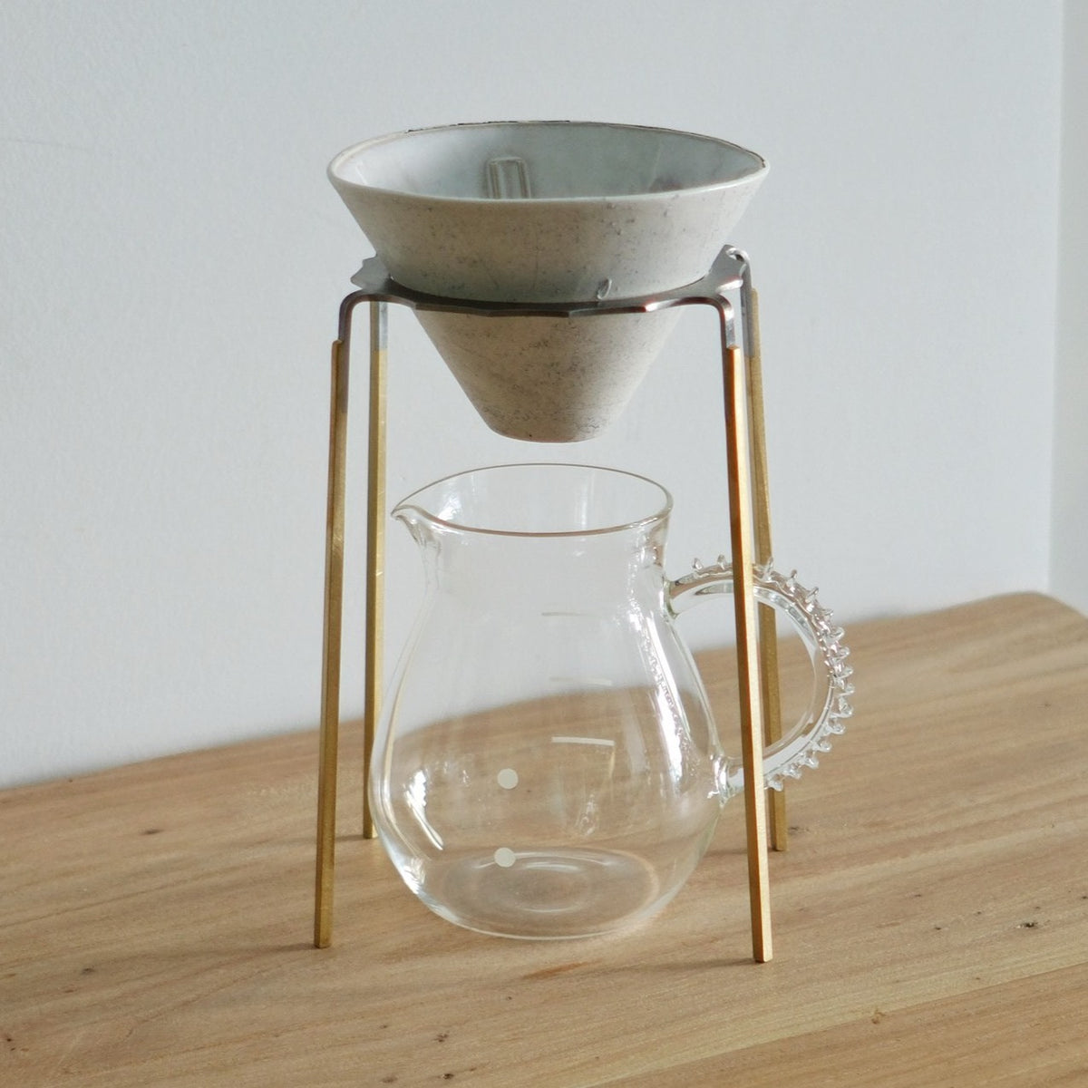 Cream ceramic coffee cone held in a slim brass stand above a clear glass serving pitcher.