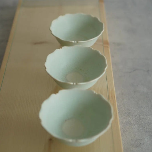 Three pale jade-green bowls with scalloped, petal-like rims, lined up on a light wooden surface.