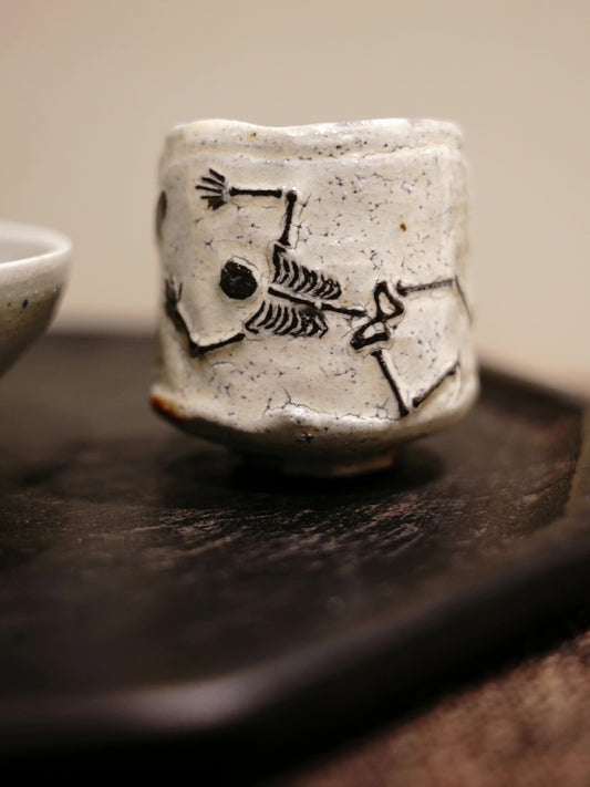 Cream speckled sake cup drawn with a simple black skeleton figure stretching around its side.