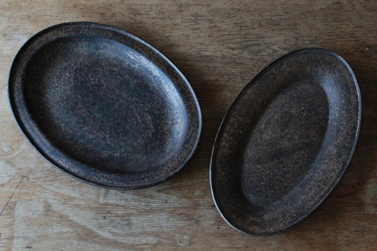 Two oval dishes in deep speckled black lacquer, one upright and one lying flat on a pale wooden board.