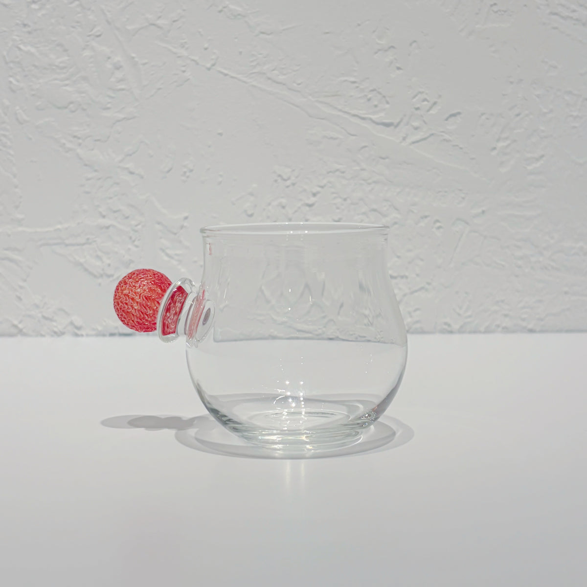 Rounded clear glass cup on a white surface, a small red glass ball resting against its side.
