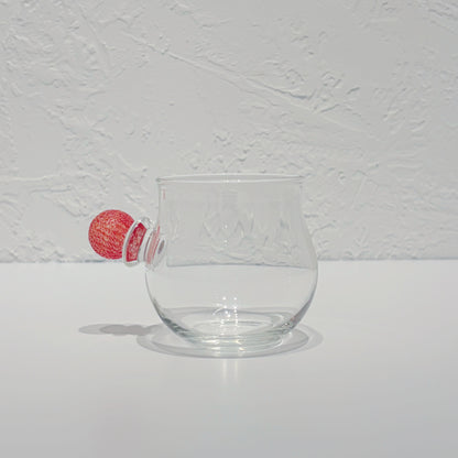 Rounded clear glass cup on a white surface, a small red glass ball resting against its side.
