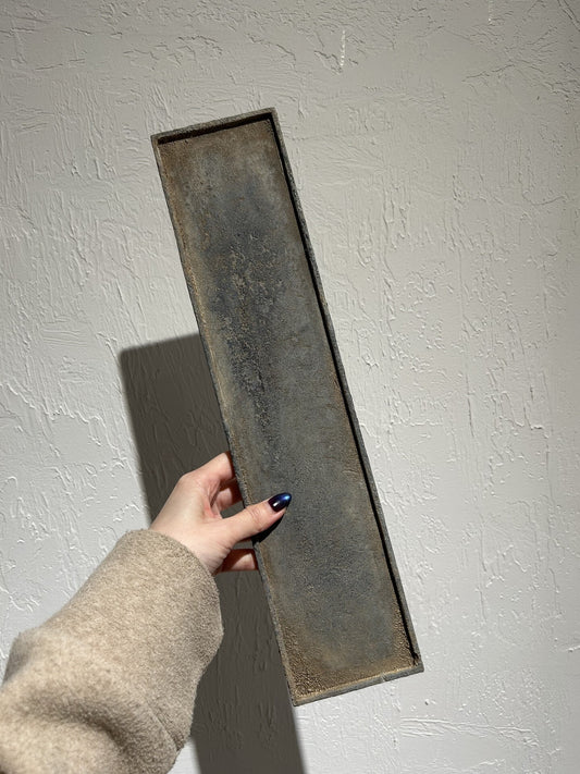 Long angular dish with an iron-corroded finish in mottled brown and grey, held up against a white wall.