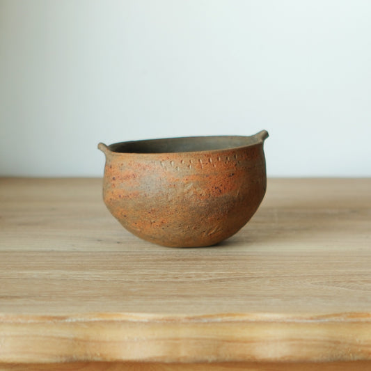 Two-handled bowl in wood-fired sandy brown with small lugs at the rim, on a wooden table.