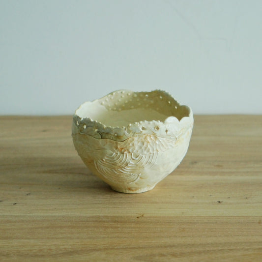 Cream tea bowl with a frilled lace-like rim and a finely combed surface, on a light wooden table.
