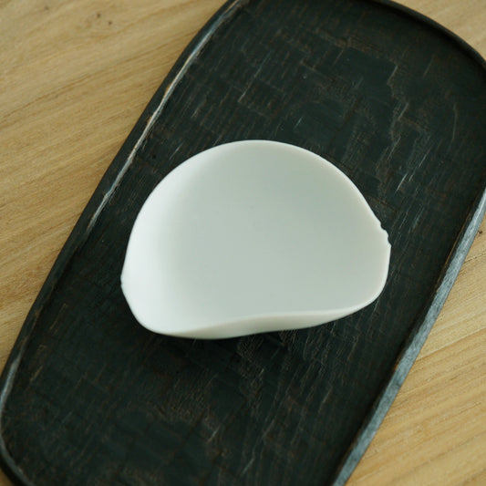 Curved white porcelain tea leaf holder shaped like a folded shell, on a dark board.