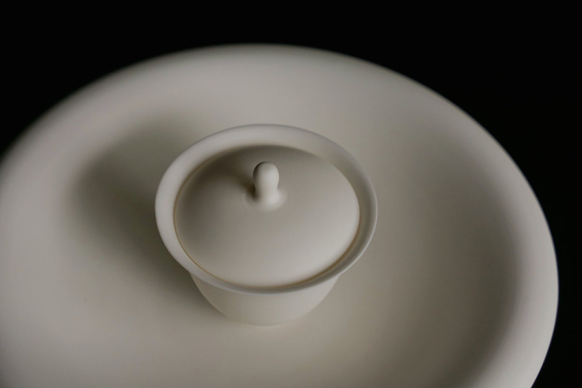 White porcelain lidded bowl with a small pointed knob, sitting on a matching white saucer.