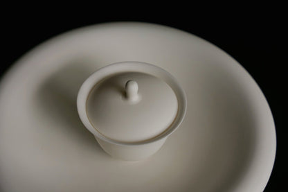 White porcelain lidded bowl with a small pointed knob, sitting on a matching white saucer.