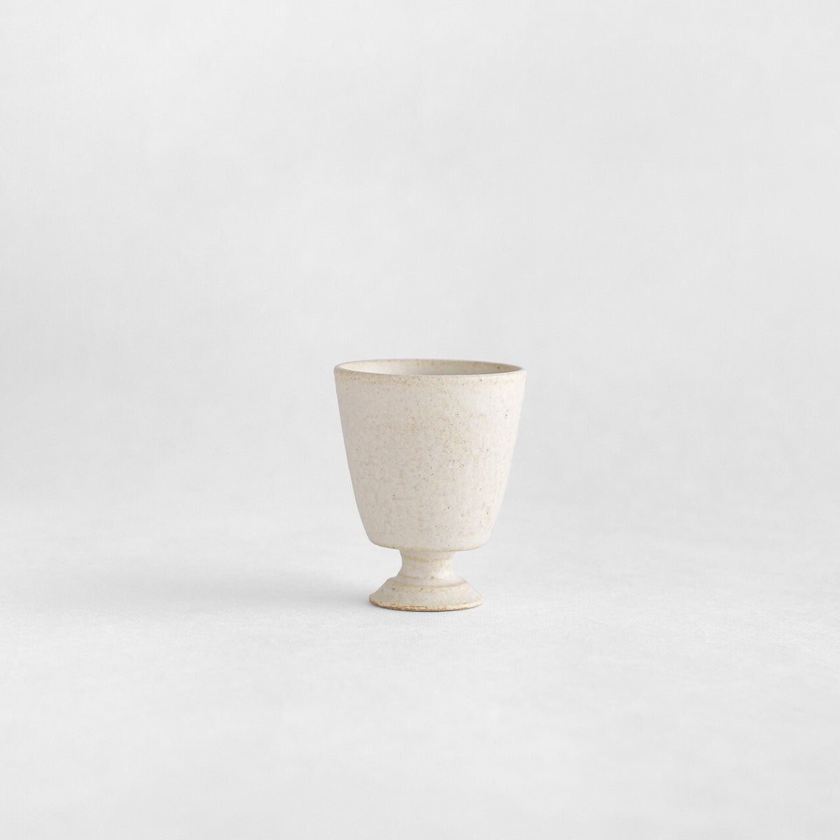 Small cream stemmed tea cup with a finely speckled matte surface, standing alone on white.
