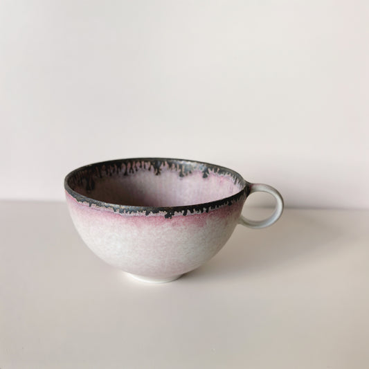 Pale pink cup with a dark speckled rim running into the white body, with a small loop handle.