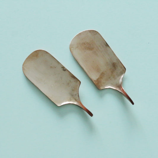 Two broad copper scoops with slim tapering handles, laid on a pale blue ground.