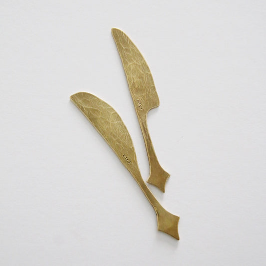 Two brass butter knives with textured blades and small forked tips at the handle ends.