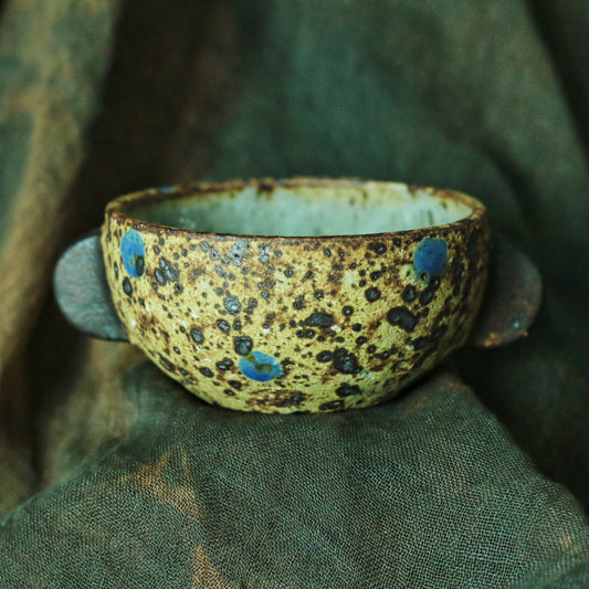 Two-handled bowl in bright speckled yellow with blue spots, resting on dark green cloth.