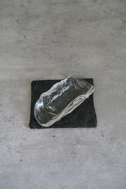 Long curved clear glass tea scoop resting on a small dark stone slab.