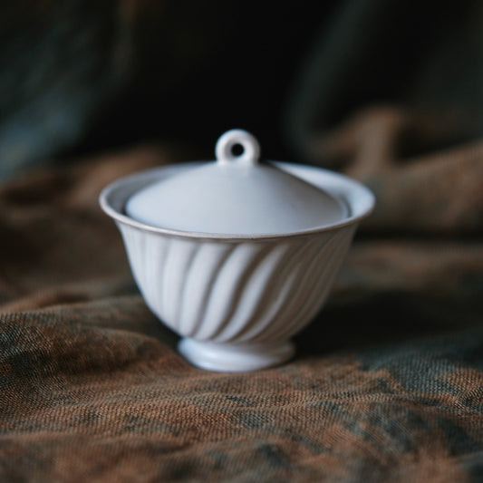 Fluted pale grey lidded bowl with a small ring knob, on soft brown cloth.