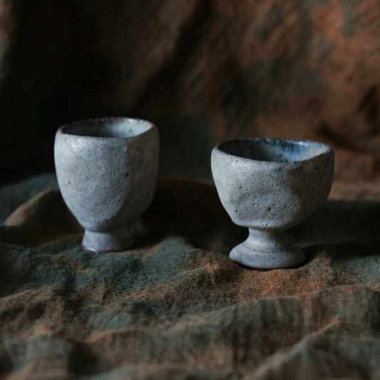 Two small footed cups in speckled blue-grey, standing side by side on dark cloth.