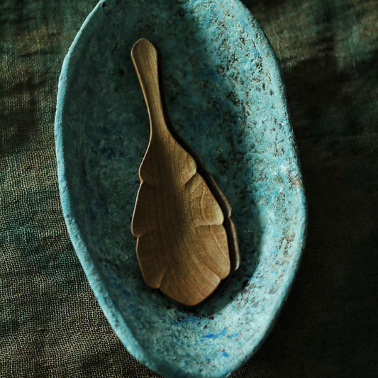 Carved wooden tea scoop with a leaf-shaped bowl, resting in a turquoise speckled dish.
