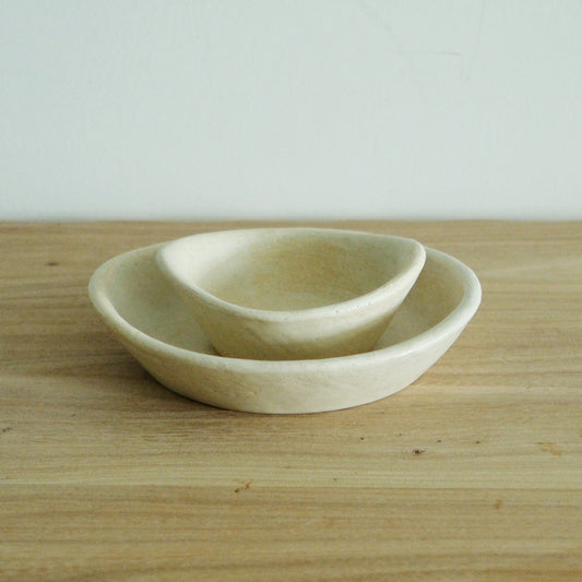 Two small oval bowls in cream and pale yellow, one resting inside the other on a wooden table.