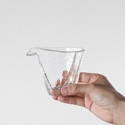 Angular clear glass pouring bowl with a sharp drawn spout, held in a hand against white.