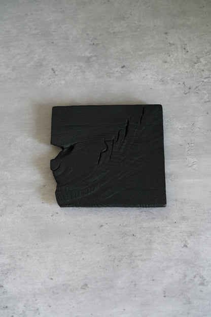 Small square coaster of charred wood in matte black, one edge cut in a jagged step.