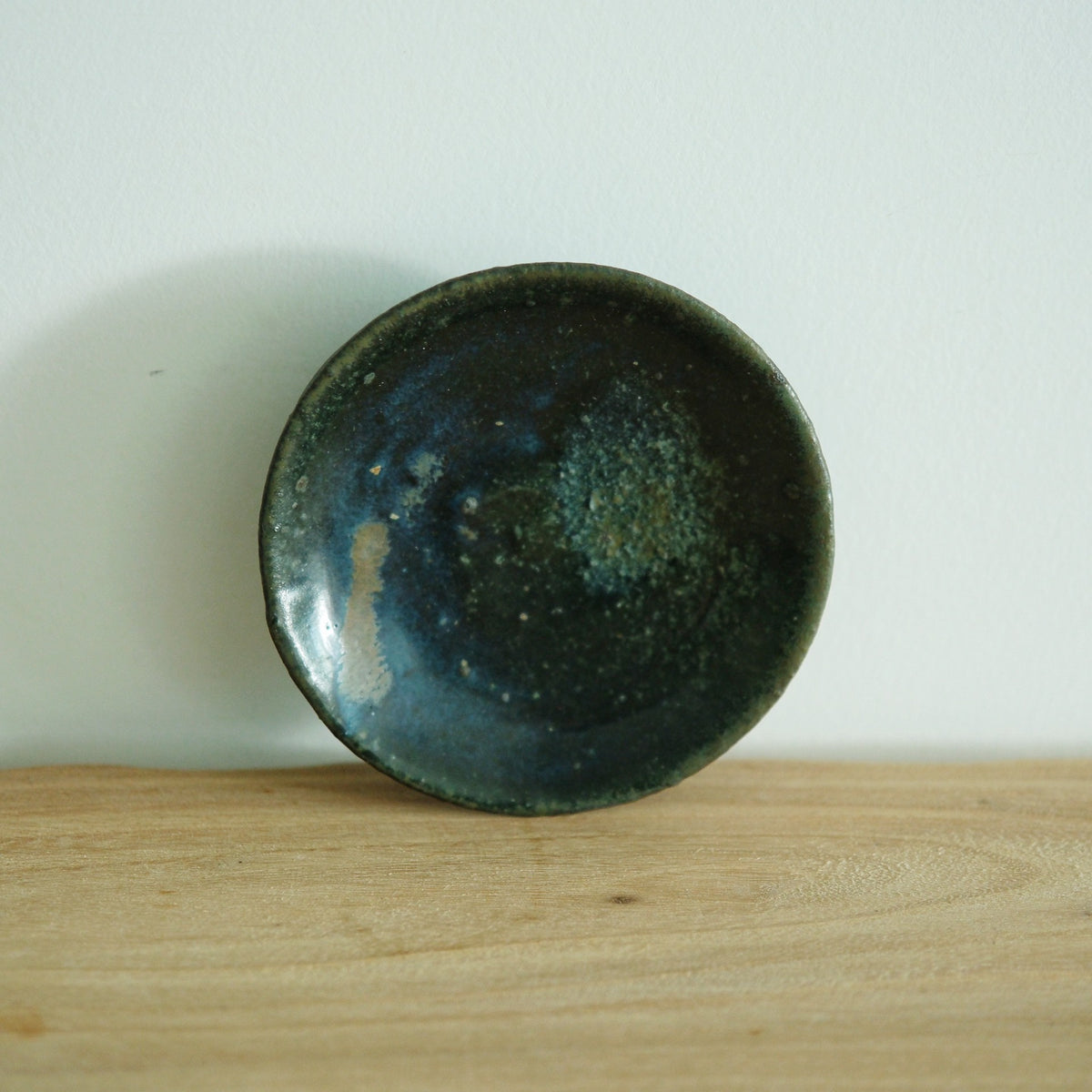 Small round dish glazed in deep teal and black, standing upright on a wooden table.