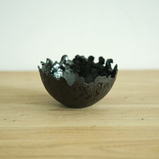 Dark bowl with a jagged torn rim and a glossy green-black glaze, on a light wooden table.