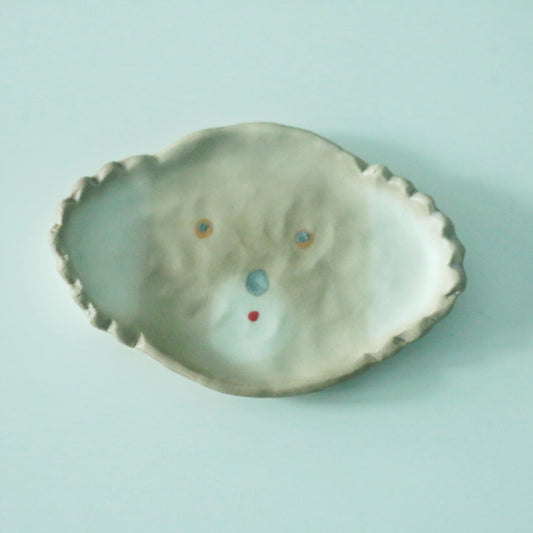 Small pale green dish with a frilled edge, painted with a simple face in soft blue and red dots.