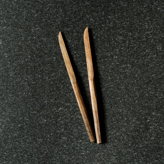 Two slender carved wooden sweet knives with pointed tips, lying on dark speckled stone.