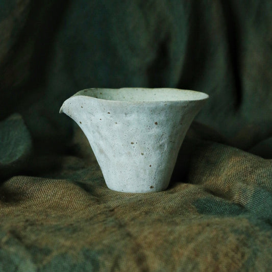 Small sand-coloured pouring vessel flaring sharply at the rim, on rumpled green cloth.