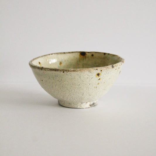 Cream rice bowl on a dark clay foot, its speckled slip broken by a few iron spots.