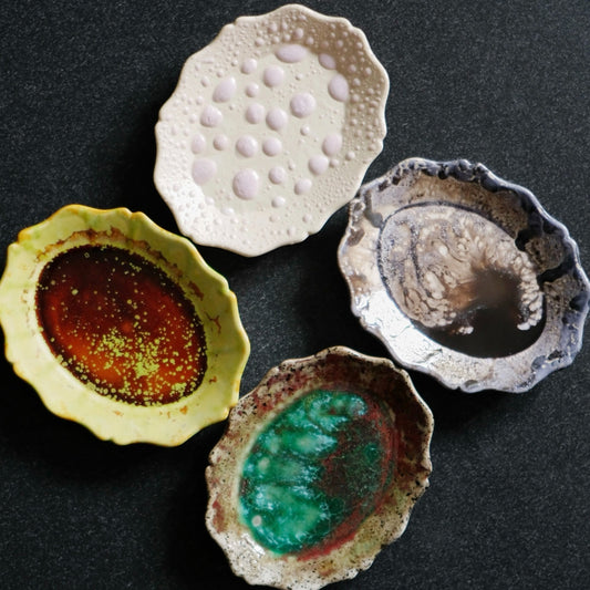 Four small scalloped dishes with pooled glazes in amber, jade green, silver and white, on black.