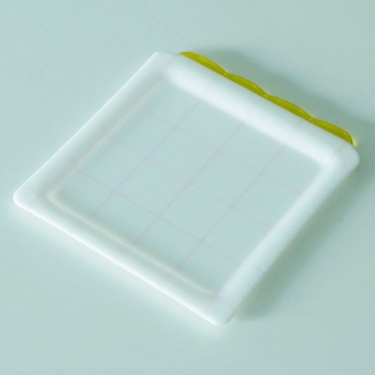 Square milky white glass plate with a faint check grid and a bright yellow stripe along one edge.