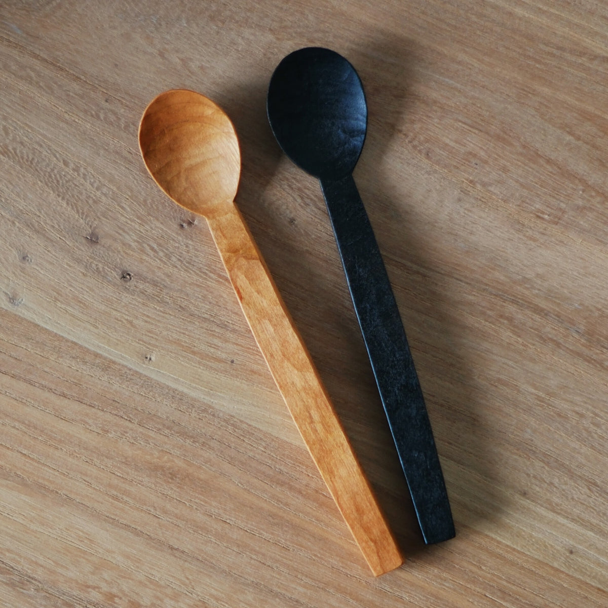 Two carved wooden spoons side by side, one pale honey and one finished in black, on a wooden table.