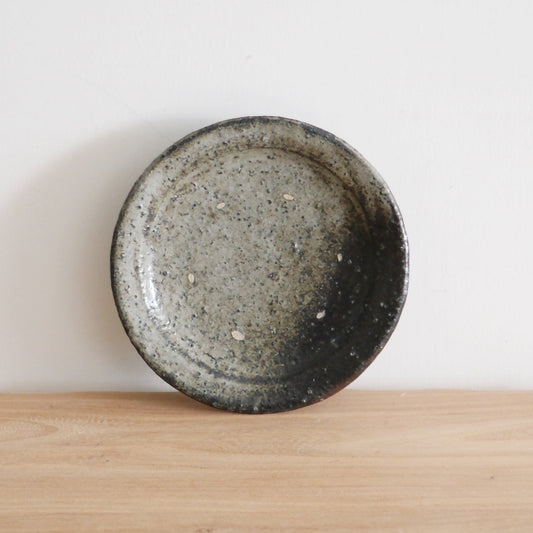 Round wood-fired plate in mottled grey and black with a heavily speckled surface, on a wooden table.