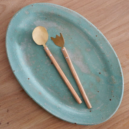 A brass spoon and a matching brass fork with wooden handles, laid across a speckled blue plate.