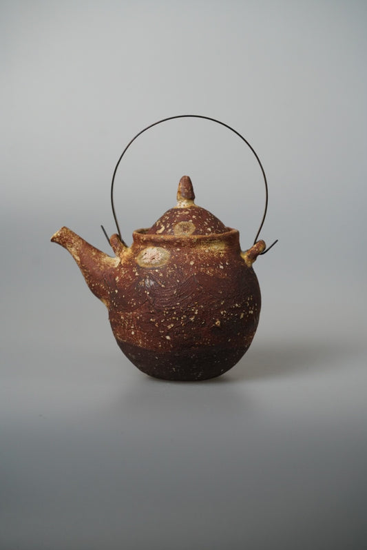 Round wood-fired teapot in mottled rust brown with a tall arched wire handle and a knopped lid.