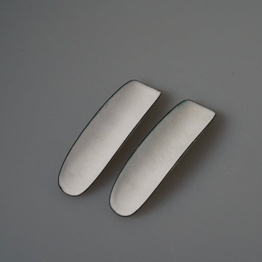 Two long narrow enamel tea scoops in soft white with dark rims, side by side on grey.