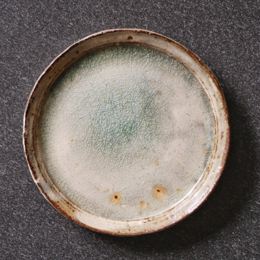Flat round plate with a soft pale green glazed face and a warm rust-brown rim, on grey cloth.