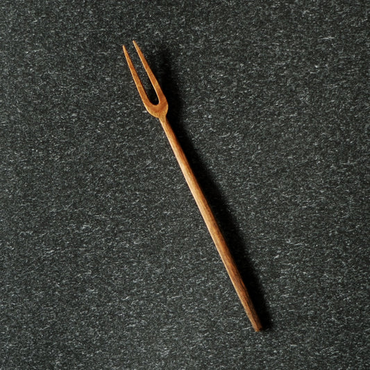 Slim two-pronged wooden fruit fork with a long tapered handle, on dark speckled stone.