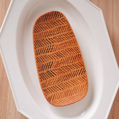 Small oval wooden tray carved in fine chevron rows, resting inside a white plate.