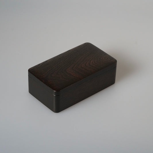 Rectangular lidded wooden box in dark lacquer, its grain showing faintly through the surface.