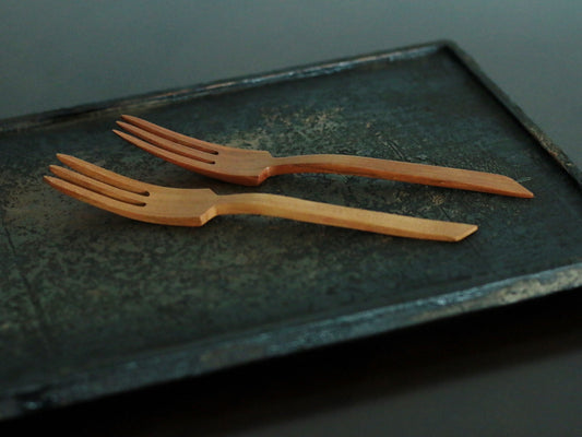 Two slim carved wooden fruit forks lying side by side on a dark metal tray.