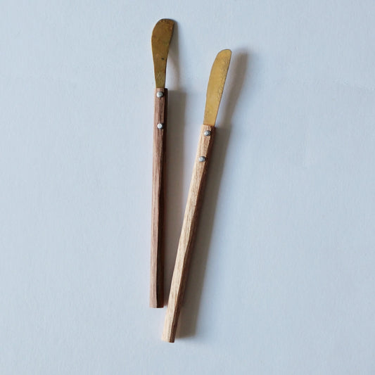 Two brass spreading knives with narrow blades and slim wooden handles, on a pale ground.