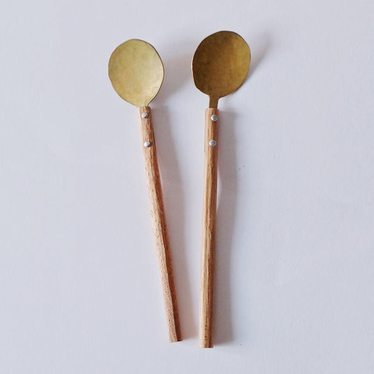 Two brass dessert spoons with long tapered wooden handles, on a pale ground.