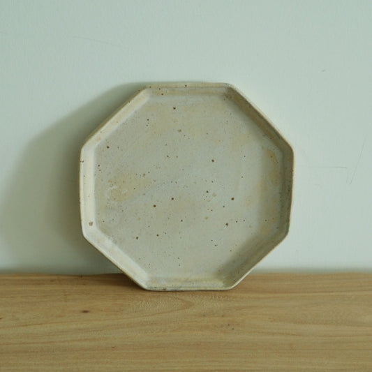 Octagonal plate in speckled cream with a wide flat rim, standing upright on a wooden shelf.