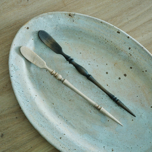 A slim tea spoon and a pointed tea needle with twisted dark handles, on a speckled green plate.
