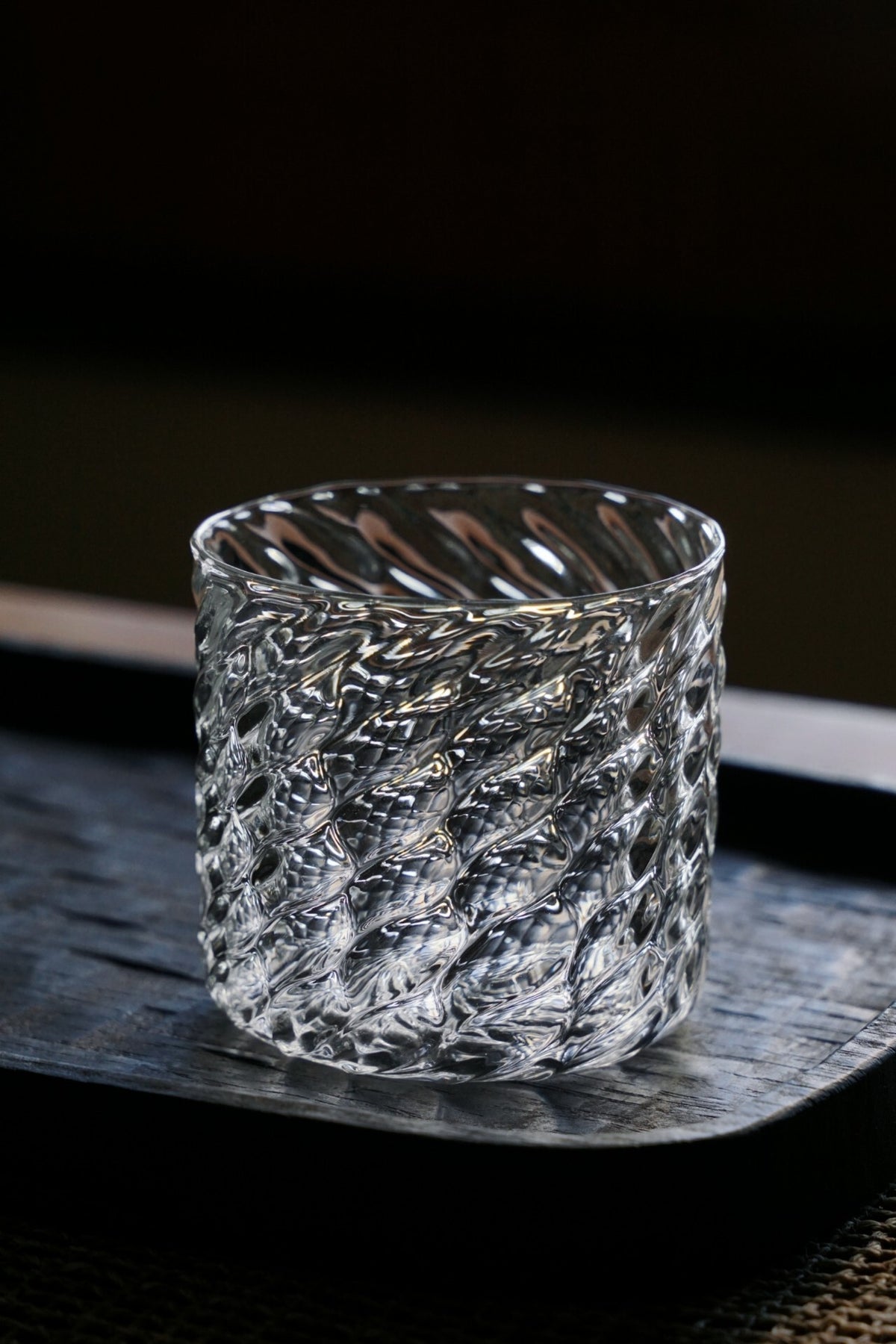 Straight-sided clear glass cup covered in a fine diagonal scale pattern, on a dark tray.